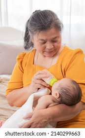 Asian Grandmother Feeding Newborn Baby In Bedroom. Senior Women Taking Care Adorable Infant At Home. Mother Enjoying Feeding Toddler And Feeling Proud. Little Child Hungry In Morning.