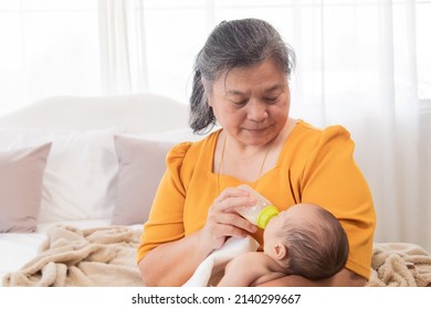 Asian Grandmother Feeding Newborn Baby In Bedroom. Senior Women Taking Care Adorable Infant At Home. Mother Enjoying Feeding Toddler And Feeling Proud. Little Child Hungry In Morning.