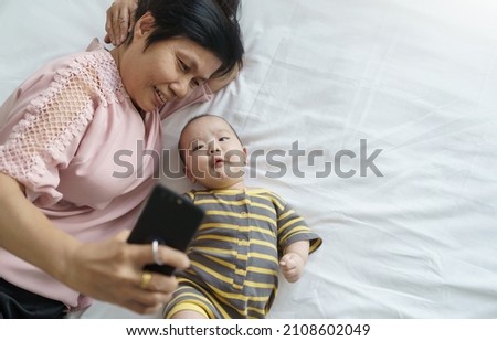 Asian Grandmother and little baby boy lying on bed using smartphone at home.