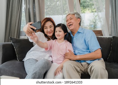 Asian Grandparents Selfie With Granddaughter At Home. Senior Chinese, Grandpa And Grandma Happy Spend Family Time Relax Using Mobile Phone With Young Girl Kid Lying On Sofa In Living Room Concept.