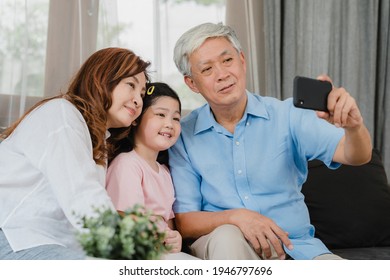 Asian Grandparents Selfie With Granddaughter At Home. Senior Chinese, Grandpa And Grandma Happy Spend Family Time Relax Using Mobile Phone With Young Girl Kid Lying On Sofa In Living Room Concept.