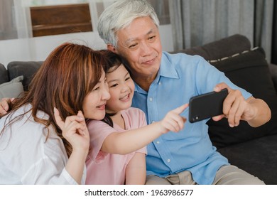 Asian Grandparents Selfie With Granddaughter At Home. Senior Chinese, Grandpa And Grandma Happy Spend Family Time Relax Using Mobile Phone With Young Girl Kid Lying On Sofa In Living Room Concept.