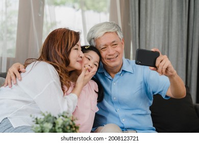 Asian Grandparents Selfie With Granddaughter At Home. Senior Chinese, Grandpa And Grandma Happy Spend Family Time Relax Using Mobile Phone With Young Girl Kid Lying On Sofa In Living Room Concept.