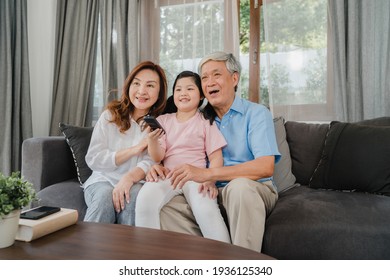 Asian Grandparents Watch TV With Granddaughter At Home. Senior Chinese, Grandfather And Grandmother Happy Using Family Time Relax With Young Girl Kid Lying On Sofa In Living Room Concept.