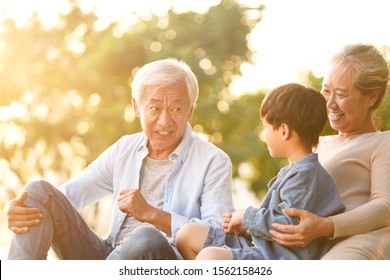 Asian Grandson, Grandfather And Grandmother Sitting Chatting On Grass Outdoors In Park At Dusk