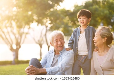 Asian Grandson, Grandfather And Grandmother Sitting Chatting On Grass Outdoors In Park At Dusk