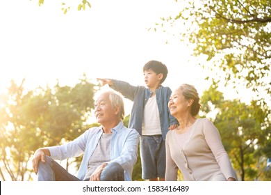 Asian Grandson, Grandfather And Grandmother Sitting Chatting On Grass Outdoors In Park At Dusk