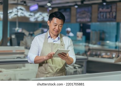 Asian Grocery Store Manager Salesman In Apron Using Digital Tablet Counting Goods In Supermarket. Male Worker Employee Doing Inspection Of Products Regular Inventory To Control Goods Online Order