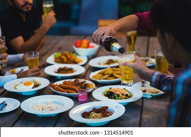 Asian Group Eating And Drinking Cold Beer Outside The House At Night, Having Fun Talking