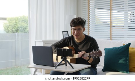 Asian Guitarist Livestream Video Or Virtual Video Tutorial At Home,artist Musician Blogger Or Vlogger Sit On Sofa In The Living Room Explaining To Play Guitar,online Learning During Quarantine Concept