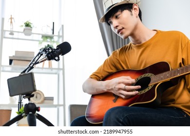 Asian Guitarist Livestream Virtual Video Tutorial At Home,artist Musician Blogger Sit On Sofa In The Living Room . Happy Young Man Singer Use Music Instrument, Compose Song On Cellphone.