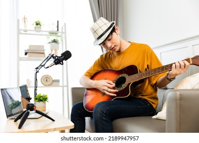 Asian Guitarist Livestream Virtual Video Tutorial At Home,artist Musician Blogger Sit On Sofa In The Living Room . Happy Young Man Singer Use Music Instrument, Compose Song On Cellphone.