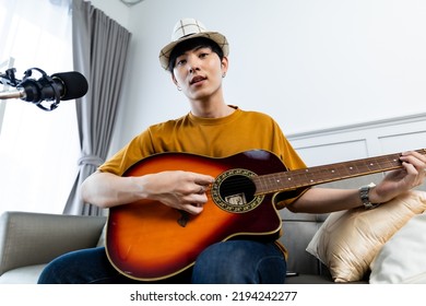 Asian guitarist livestream virtual video tutorial at home,artist musician blogger sit on sofa in the living room . Happy young man singer use music instrument, compose song on cellphone.