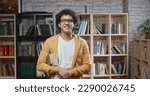 Asian guy with curly hair is standing between bookshelves of library holding books in hands, looking at camera and smiling - portrait closeup. education, student lifestyle concept 