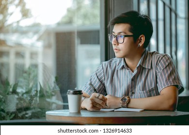 Asian guy with glasses sitting and writing something with thought.