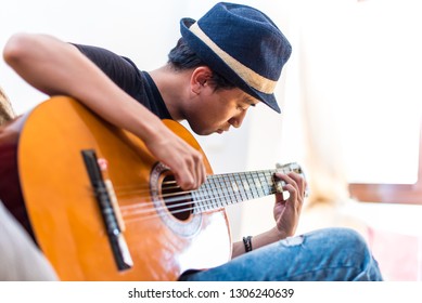 Asian Guy With A Hat Playing Guitar At Home. Man Playing Guitar  In His House. 