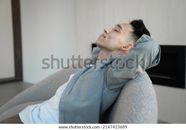Asian guy leans on armchair back nodding head while\
listening to dynamic music. Young man relaxes at home on blurred\
background close side\
close