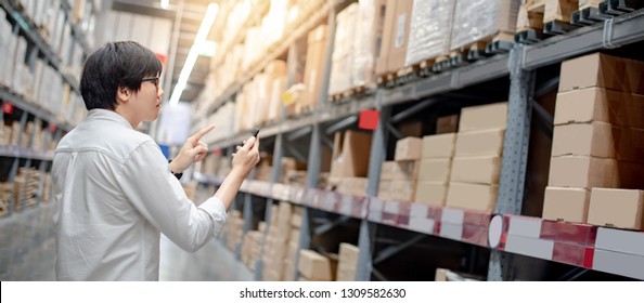 Asian guy shopper standing in warehouse inventory aisle checking shopping list on smartphone. Buying or purchasing factory goods. Shopaholic concept