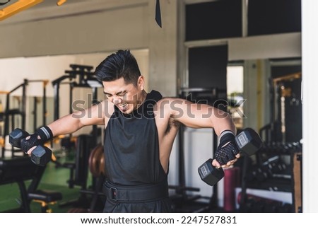 An asian guy struggling to do a set of standing dumbbell side lateral raises at the gym. Enduring the last reps, feeling muscle burn. Weight training at the gym.