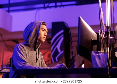 Asian hacker breaking law while doing data mining using malicious software. Young man in hood hacking into computer system while cracking password in abandoned warehouse at night