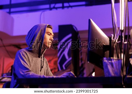 Asian hacker breaking law while doing data mining using malicious software. Young man in hood hacking into computer system while cracking password in abandoned warehouse at night
