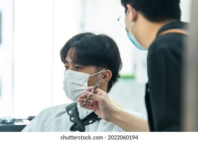 Asian Hairdresser Barber Wear Mask, Cut Hair Of Attractive Man In Shop. Handsome Young Man Customer In Modern Salon Barbershop, Enjoy Getting Haircut Service By Stylist Man During Covid 19 Pandemic.