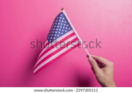 Asian hand holding the American flag with stars and stripes design, set against a pink background. Symbolizes U.S. pride, patriotism, democracy, holidays, and national identity or unity.