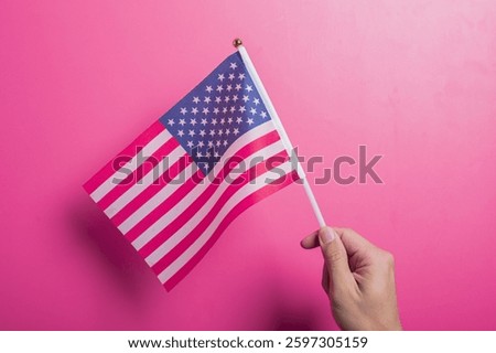 Asian hand holding the American flag with stars and stripes design, set against a pink background. Symbolizes U.S. pride, patriotism, democracy, holidays, and national identity or unity.