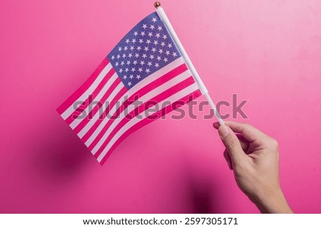 Asian hand holding the American flag with stars and stripes design, set against a pink background. Symbolizes U.S. pride, patriotism, democracy, holidays, and national identity or unity.
