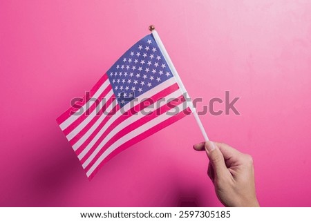 Asian hand holding the American flag with stars and stripes design, set against a pink background. Symbolizes U.S. pride, patriotism, democracy, holidays, and national identity or unity.