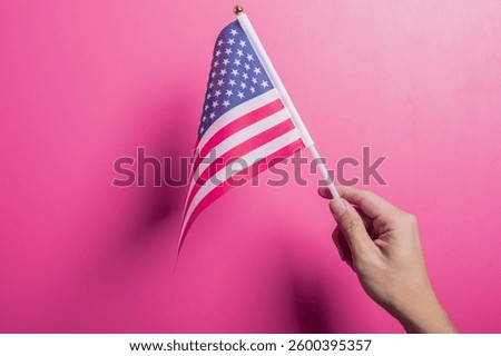 Asian hand holding the American flag with stars and stripes design, set against a pink background. Symbolizes U.S. pride, patriotism, democracy, holidays, and national identity or unity.