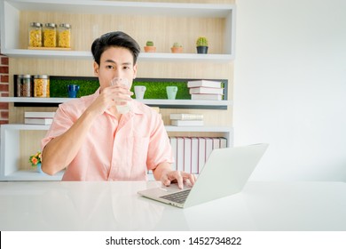 A Asian Handsome Businessman 24s Drink Milk And Working With Laptop On Desk In Workplace. A Handsome 24 Year Old Asian Male Is Drinking Milk And  Use A Laptop On The Table In The House. Work Form Home