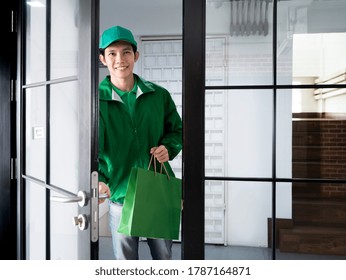 Asian Handsome Delivery Man Holding Shopping Paper Bag And Opening Door To Send Food Or Product Order By Express Service To Customer While Wearing Green Jacket Uniform And Cap At Apartment Room