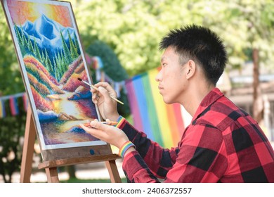 Asian handsome gay wears rainbow wristband and reparing his art mountain drawing in the park, concept for lifestyle and recreational activity of LGBT people around the world, new edited.