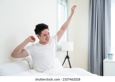 Asian handsome male in pajamas wake up in the morning with happiness. Attractive young man smiling, feeling happy and relax then stretching body after getting up from sleep on bed in bedroom at home.