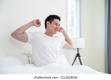 Asian Handsome Male In Pajamas Wake Up In The Morning With Happiness. Attractive Young Man Smiling, Feeling Happy And Relax Then Stretching Body After Getting Up From Sleep On Bed In Bedroom At Home.