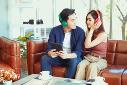 Asian Handsome Man And Beautiful Woman Lovely Couple Choose Music On Red Tablet Device For Listen On Headphone Setting On Brown Leather Sofa In Home With Cute Balloon Decoration For Winter Party.