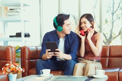 Asian Handsome Man And Beautiful Woman Lovely Couple Choose Music On Red Tablet Device For Listen On Headphone Setting On Brown Leather Sofa In Home With Cute Balloon Decoration For Winter Party.