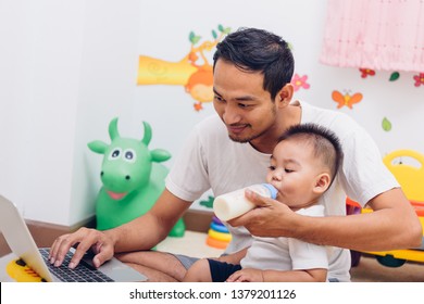 Asian Handsome Man Father, But He's Acting Mom Parent He Is Feeding Milk His Son Baby 1 Year Old While Working On Laptop Computer At Home Office, Work From Home With Family Concept