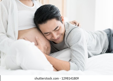 Asian Handsome man is listening to his Asian beautiful pregnant wife's tummy and smiling. Family love concept.