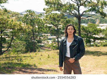 An asian handsome man with long hair in a black leather jacket stands confidently on a dirt road, surrounded by nature, with a clear sky above him. - Powered by Shutterstock