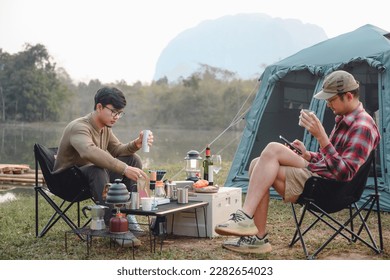 Asian handsome man Sitting on a camp chair on the lawn relaxing on vacation. Two male tourists drinking drip coffee in a natural forest Likes to travel and go camping