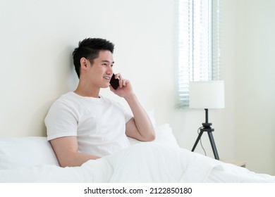 Asian Handsome Man Talk On Mobile Phone Call On Bed At Home In Morning. Happy Attractive Male Lying Down In Bedroom Enjoy Holiday Weekend, Using Smartphone Communication With Technology In House.