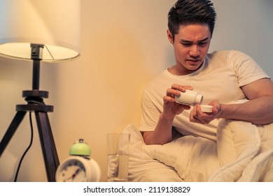 Asian Handsome Sick Male In Pajamas Take Medicine In Dark Night Room. Attractive Young Man Patient Take Drugs From Bottle And Rest While Lying Down On Bed In Bedroom. Health Care And Medical Concept.