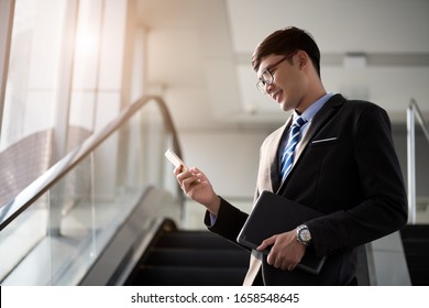 A asian handsome young businessman holding his laptop tablet while using his phone and standing on the escalator. Business concept. - Powered by Shutterstock