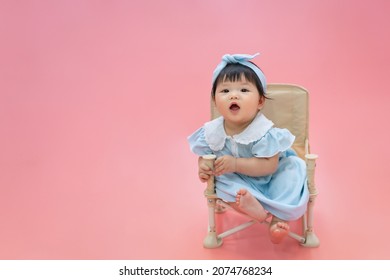 Asian Happy Baby Smiling And Sitting On A Chair In Pink Color Background. Cute 6 Months Baby Wearing Blue Cloth With Copy Space Use As Concept Of Valentine, Love, Baby Or Kid Department In Hospital.