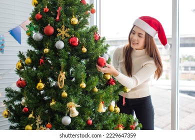 Asian happy cheerful female housewife in sweater and red Santa Claus hat standing smiling holding hanging shiny glitter golden ball decorating pine tree celebrating Christmas eve in home living room. - Powered by Shutterstock