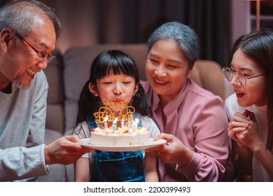 Asian happy family of little girl blowing out candles on cake. Celebrate brithday anniversary party with  Grandparents and mother on table at night in living room. Kid girl having happiness lifestyle.
