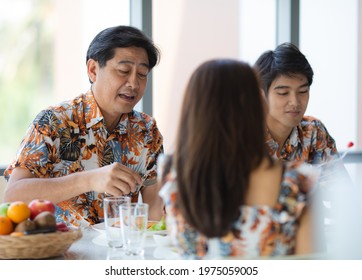 Asian Happy Family Middle Aged Father And Teen Son Wears Colorful Team Uniform Shirt Talking Funny Together On Dining Table Eat Lunch Behind Mother Sitting On Opposite Chair In Blurred Foreground.