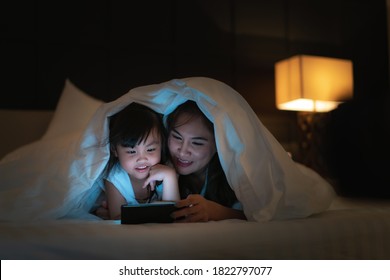 Asian happy family mother and little daughter watching movie or cartoon in smartphone together and blanket cover their head in bed at night at home - Powered by Shutterstock